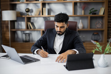 Fototapeta premium Young bearded male freelancer in business suit signing papers while sitting at table with laptop and tablet in stylish office. Remotely work.