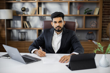 Fototapeta na wymiar Young bearded male freelancer in business suit signing papers while sitting at table with laptop and tablet in stylish office. Remotely work.