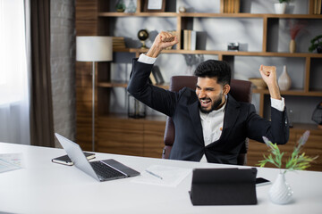 Naklejka premium Excited screaming young bearded business man or entrepreneur working with laptop computer and tablet while triumphing with raised hands because successful business deal.