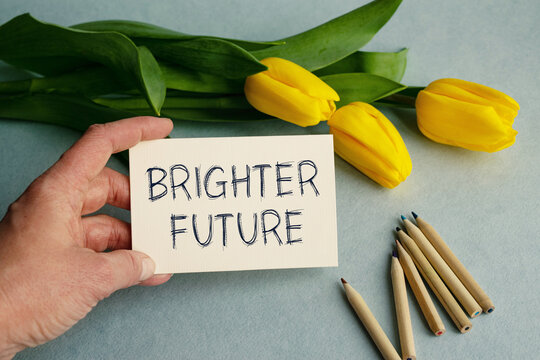 The hand of a young girl close-up holds a card with the inscription BRIGHTER FUTURE on a light blue background along with yellow tulip flowers. View from above. Conceptual image.