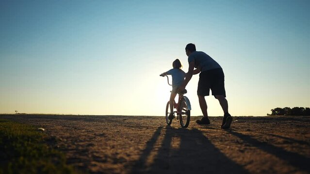 dad teaches daughter to ride a bike. happy family childhood dream concept. father and little daughter learn to ride a bike silhouette in the park. happy family goes in for sports outdoors sunlight