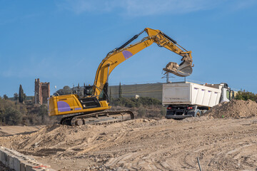 Large yellow excavator depositing sand in a truck bed at a large construction site with disturbed earth