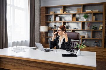 Corporate communication. Caucasian young beautiful businesswoman talking on mobile phone while working on laptop in modern office. Free Space