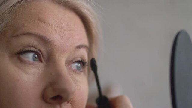 portrait of a caucasian blonde woman 40-45 years old painting eyelashes in front of a deorge mirror in her hands, selective focus. a mature woman paints eyelashes. aged facial skin care
