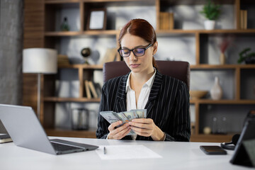 Fototapeta premium Rich business woman working on laptop counting money cash, calculate earnings income profit at office workplace. Professional manager freelancer girl. Business people. Employment, occupation