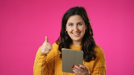 Portrait of happy smiling young woman 20s in sweater shopping on tablet computer give thumbs up gesture isolated on pink background studio. 