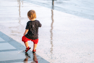 Toddler walks on a wet road in red sandals. Small child walks on a rainy day. Looks into the reflection
