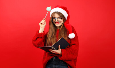 Positive young woman in a Santa hat and a notepad with wishes for the new year in her hands on a...