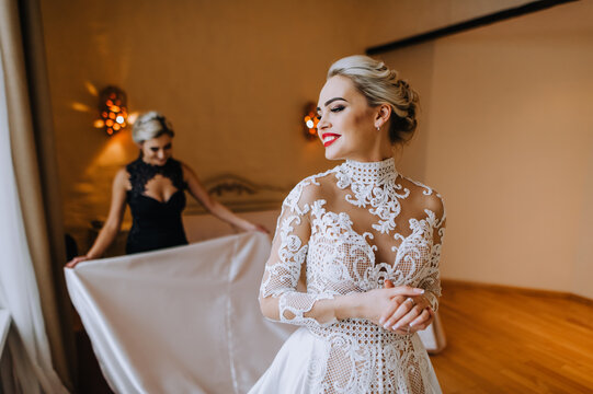 Bridesmaid helps in preparing a beautiful smiling blonde model bride in the room, hotel in the morning. Photography, wedding, portrait.