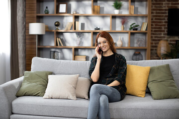 Obraz premium Happy young caucasian woman sitting on cozy sofa with modern smartphone in hands and calling to customers during break. Pretty female resting during working day at home.