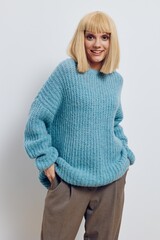 a stylish, happy blonde woman stands on a light background in a long blue sweater, smiling pleasantly and holding her hands in the pockets of brown pants
