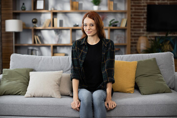Obraz premium Happy red hair caucasian woman sit rest on comfortable couch in modern living room distance dreaming. Smiling young female renter or tenant relax on sofa at home, enjoy weekend.