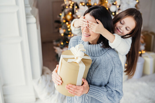 Small adorable female kid prepares surprise for her mother, closes eyes and gives wrapped present as stand at New Year background. Pretty female recieves gift from daughter. Surprisment concept