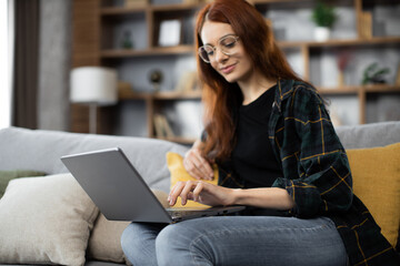 Smiling young woman using laptop, sitting on couch at home, beautiful girl having video call online in social network, having fun, watching movie, freelancer working on computer project