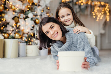 Children, family and celebration concept. Adorable female in knitted sweater holds white present box and small kid stands behind her back, have good mood before New Year`s Eve. Waiting miracle