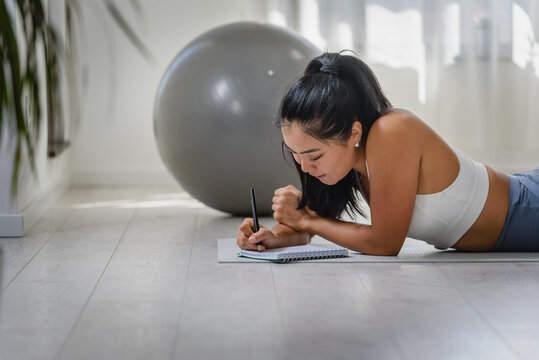 Young Asian yoga woman writing in her notebook. Sitting at home by the window after exercise. Personal growth resolutions - Powered by Adobe