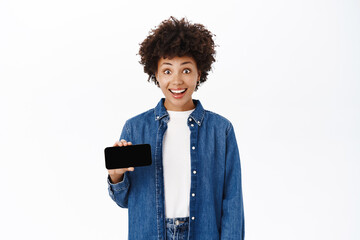 Happy young woman smiles, shows her mobile phone screen, demonstrates website or online shop on her smartphone, white background