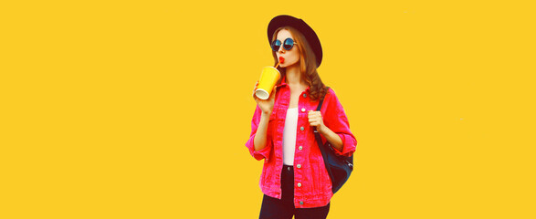 Portrait of stylish young woman drinking fresh juice wearing pink jacket, black round hat, backpack on yellow background