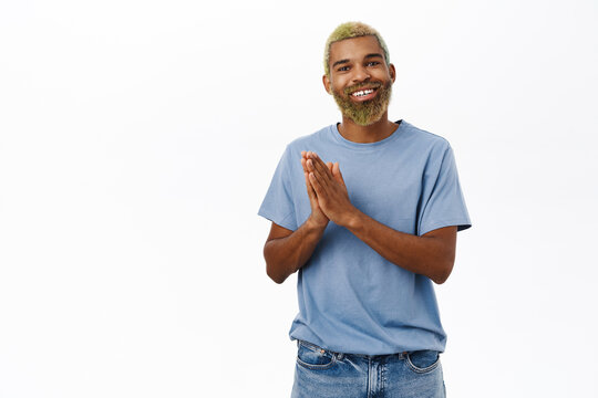 Image of young hopeful guy looking with happy smile, hold hands in pray, hopes to achieve smth, stands over white background