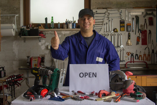 Image of a smiling handyman giving a thumbs up in his workshop with a bench full of work tools and an open sign. Starting a new craft business and new job prospects
