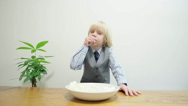 A caucasian boy is eating corn curls from the plate someone pushed in front of him on the table. A boy likes corn curls and crams them into mouth with joy