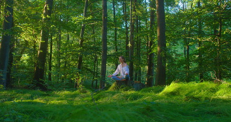 Forest green meadow glade. Treatment of mental illnes by practicing yoga in woodland. Girl sitting on stump and meditating in nature. Woman relax in lotus position and breath, closed eyes, evening.