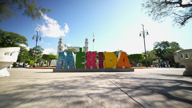 Fly over pull back from Cathedral Ildefonso to reveal the Merida sign in Merida Mexico Plaza Grande with a clear blue sky.