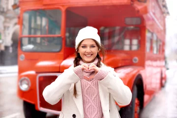 Fototapeten love winter holidays. woman makes a heart gesture on red bus city background © cenchild