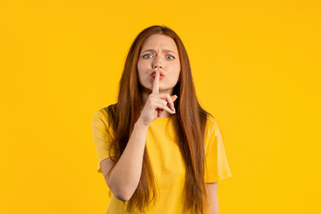 Ginger woman holding finger on lips, yellow studio background. Pretty lady with gesture of shhh, secret, silence, conspiracy, gossip concept