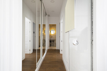 A long corridor in a house with white materials on the walls, wooden doors and a wardrobe with sliding mirror doors