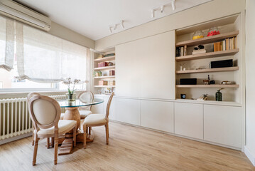 Living room decorated in a contemporary style with a round glass table, wooden chairs upholstered in smooth fabric and a bookcase with smooth furniture and wooden shelves