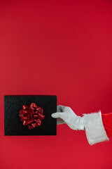 Nahaufnahme von der Hand von einem Weihnachtsmann, der ein Geschenk, vor roten Hintergrund, hält. Weihnachtlicher Hintergrund mit Platzhalter.