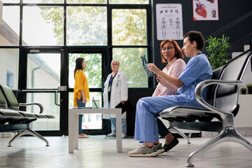 Medical nurse holding radiography scan explaining diagnosis to senior patient in hospital waiting area. Assistant discussing healthcare treatment during clinical consultation