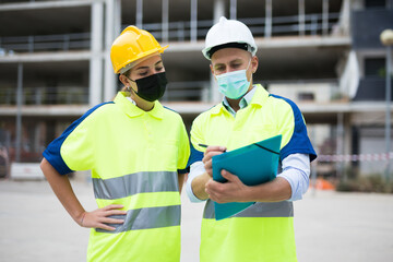 Two qualified engineers in protective masks working on a construction site during a pandemic are discussing a construction ..plan, making important work notes