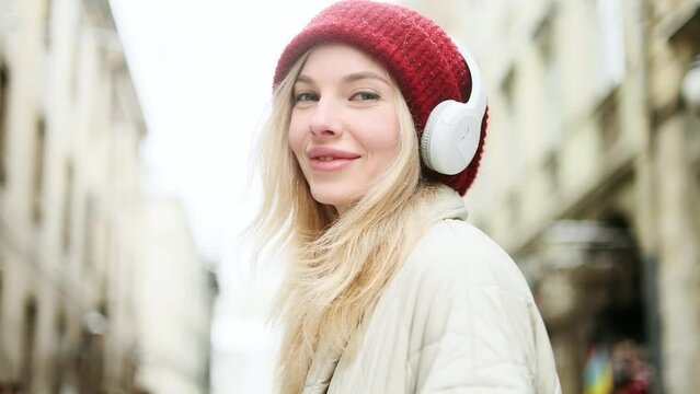 Close up portrait of charming smiling blond woman with red hat listening to music in headphones while standing on snowy winter street in city centre and looking at the camera alone