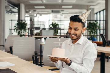 Fototapeta na wymiar Happy Hindu businessman holding cake with lightened candle in the office and looking at camera.