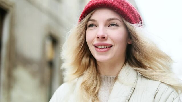 Close up portrait of charming smiling blond young woman with red hat while standing on snowy winter street in city centre and inspired looking ahead alone 