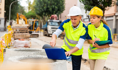 Two builders in uniform planning their work in construction plant. Man using laptop, woman holding...
