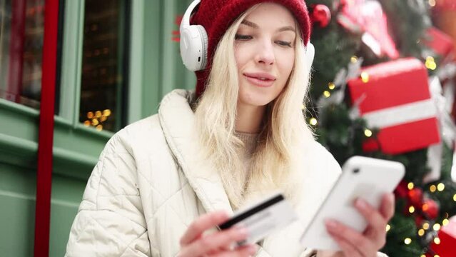 Young attractive blonde woman in red hat and headphones paying with credit card on smartphone sitting near Christmas decoration on snowy city Happy customer enjoying online shopping on holiday sales