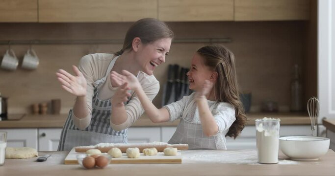 Loving cheerful mother and little daughter having fun while cooking pastries, clapping their hands sprinkling white flour, laughing feel happy, fooling around standing in cozy kitchen. Family pastime