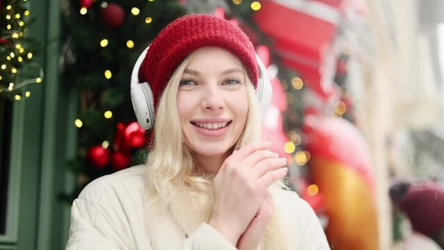 Portrait of charming blond young woman with red hat and headphones listening music while standing on snowy winter street in city centre rubbing her hands to keep warm and looking at the camera alone 