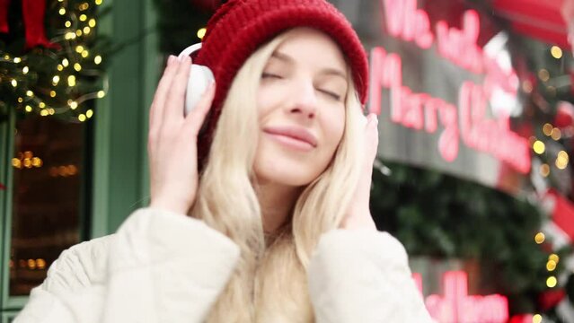 Portrait of charming blond young woman with headphones listening music while standing on snowy winter street in city centre with Christmas decoration alone 