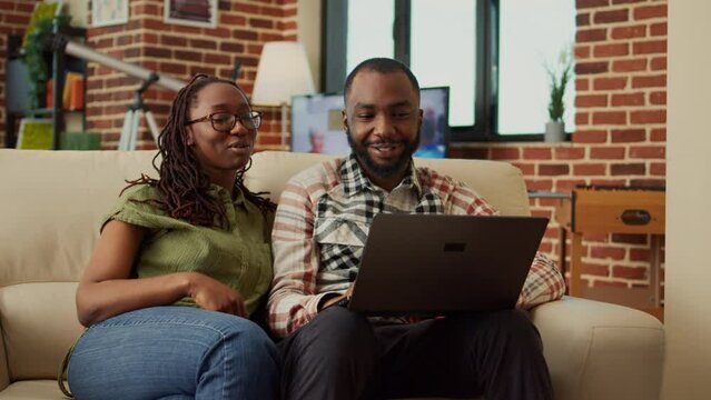 African american couple watching online videos on laptop, relaxing together on couch and having fun. Browsing internet on pc and enjoying leisure activity, laughing at home.
