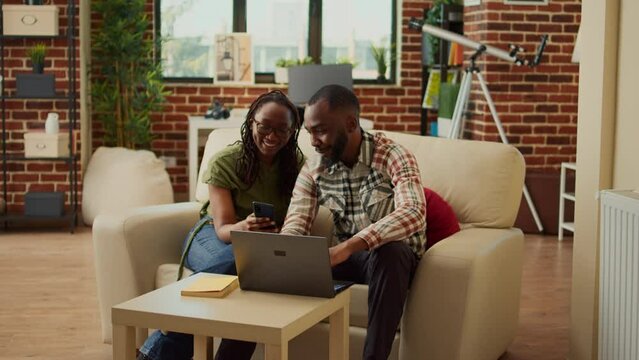 African american people relaxing at home on sofa, scrolling through social media app on smartphone. Enjoying free time leisure activity and laughing together, cozy recreation in living room.