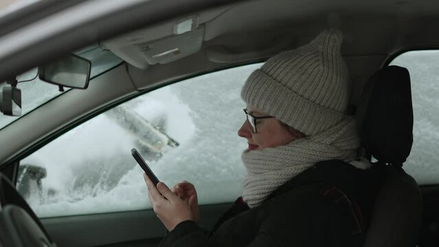 Young woman in glasses and warm clothes sits inside snow-covered car and touches smartphone screen with smile, medium shot. Unrecognizable man cleans snow from car windows with brush, slow motion.