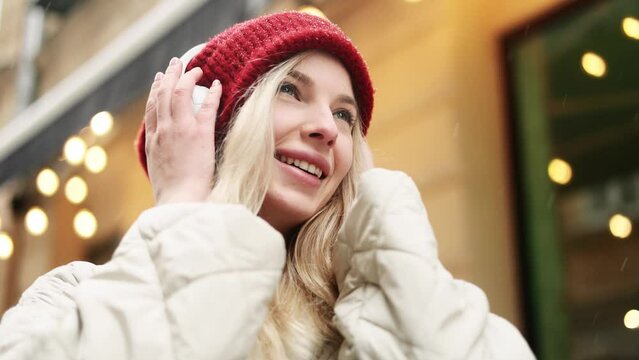 Close up portrait of charming smiling blond woman with red hat listening to music in headphones while standing on snowy winter street in city centre with light bokeh on the background alone