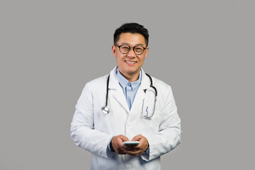 Glad adult chinese male therapist in white coat and glasses with stethoscope use smartphone for work