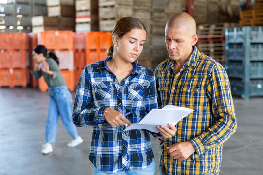 Man and young woman talking about documentation while standing in warehouse. Another woman talkin on phone in background. - Powered by Adobe