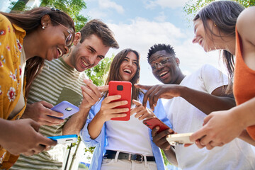 Low angle view group of young teenagers holding cell phones watching something funny on mobile screen. Surprised faces looking smartphone. Concept of technology connection community and social media 