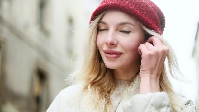 Portrait of smiling young blond woman in red hat wearing the earphones for listening music while walking down winter street in the city centre alone